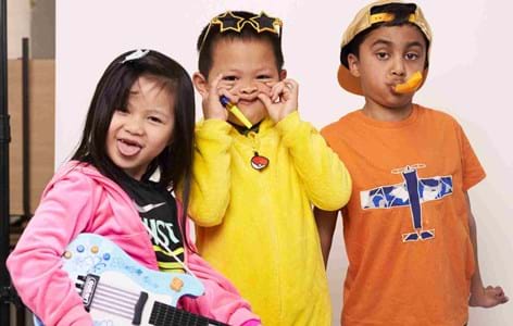 a girl in a pink hoodie with a toy guitar, a boy in yellow with star-shaped sunglasses and another boy in an orange T-shirt with a blue plane on it. The children are dressed for the Children's Mental Health Week Express Yourself campaign.