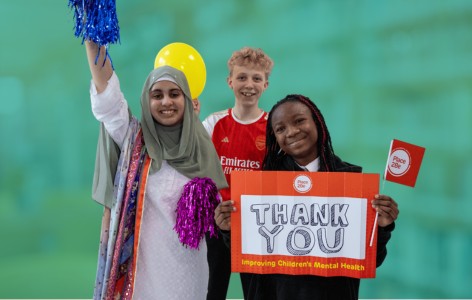 Three secondary school aged pupils all smiling in non-uniform, holding a sign saying 'Thank you', Place2Be flags and balloons.