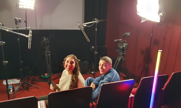 BAFTA Young Presenter Samaira Iqbal with actor Jay Lycurgo, sitting on red theatre seats, surrounded by recording equipment like cameras and microphones