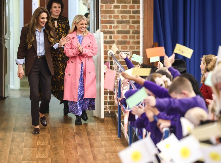 The Princess of Wales walking into a school hall at Castle Hill Academy, waving and smiling at the pupils holding Place2Be flags