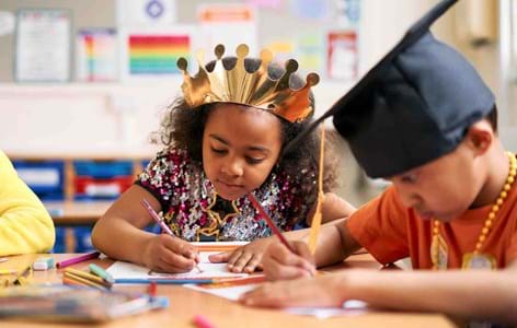 Young girl in a classroom wearing a shiny gold card crown writing with a pencil