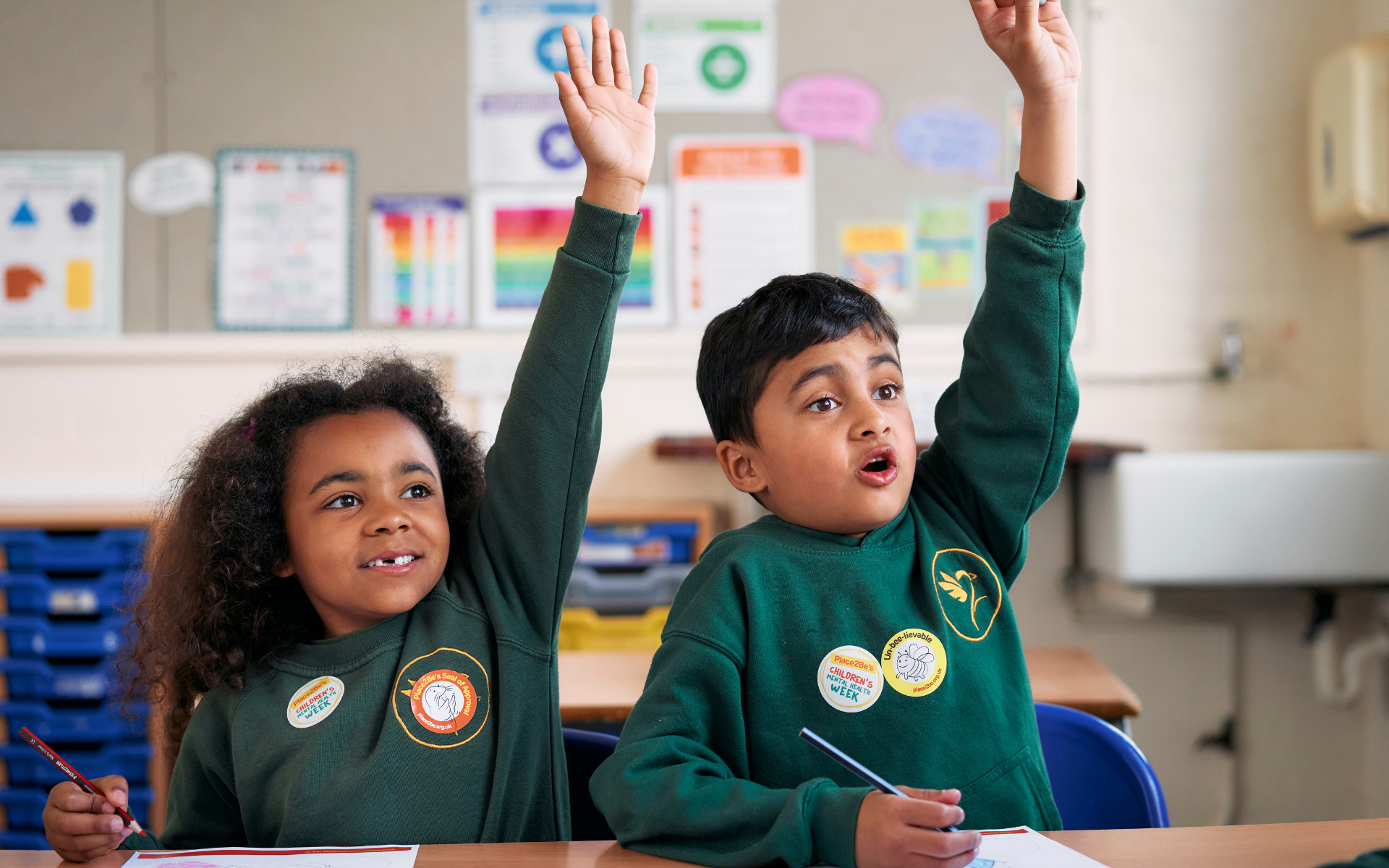 Two primary age children wearing school uniform, sat in a classroom with their hands raised. Both are wearing stickers with the Children's Mental Health Week logo on.