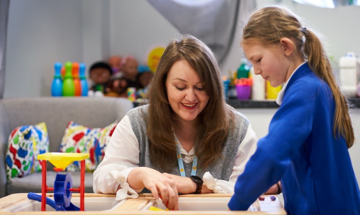 Schools Wellbeing Partnership Header Staff member with a primary school pupil in a play therapy session. They are both smiling and talking together.