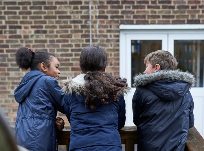 Three school pupils talking in a group leaning on a wall in the school playground. Their backs are facing the camera.