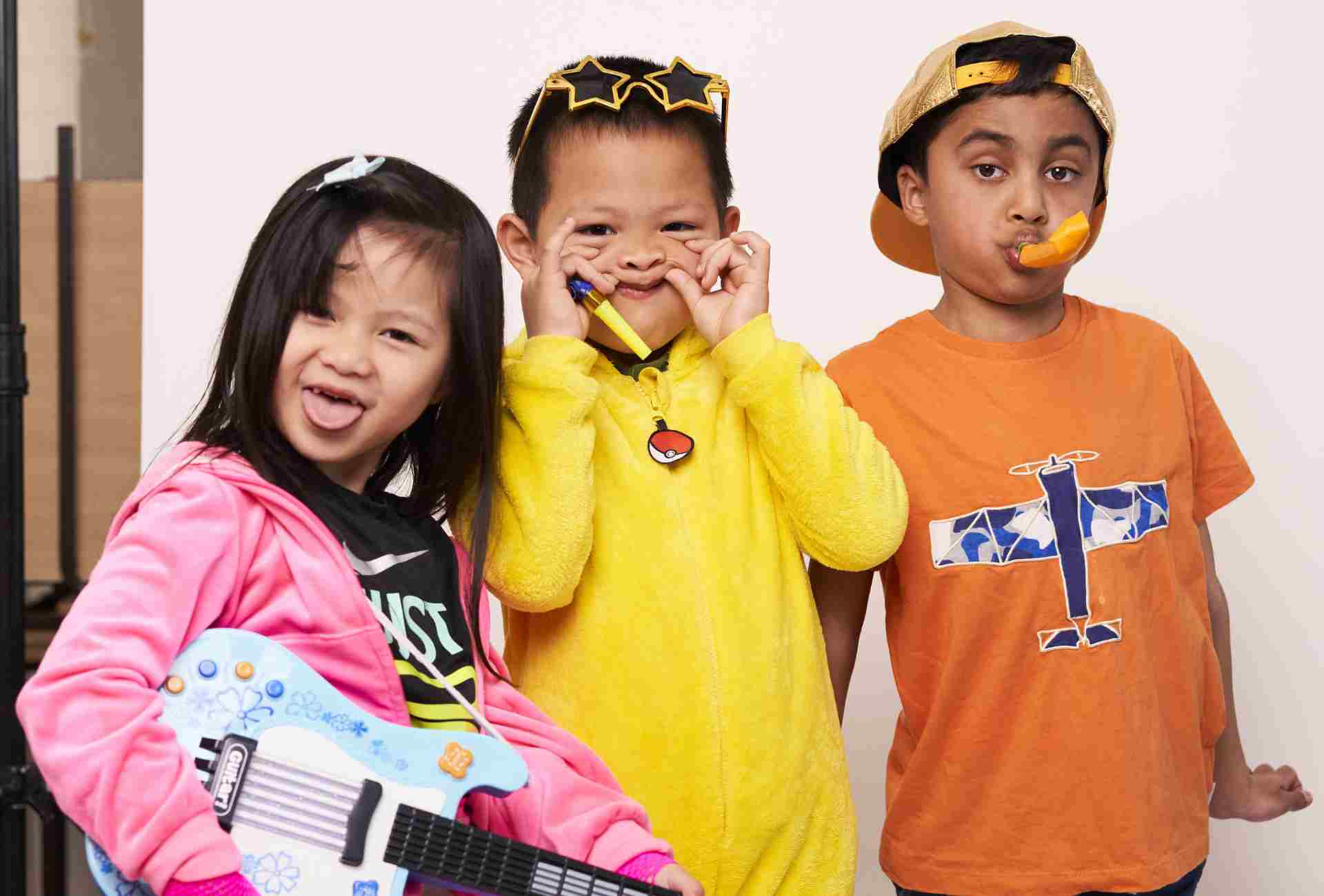 A girl in a pink hoodie with a toy guitar, a boy in yellow with star-shaped sunglasses and another boy in an orange T-shirt with a blue plane on it. The children are dressed for the Children's Mental Health Week Express Yourself campaign.