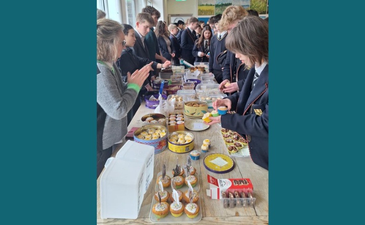 A long table filled with cupcakes, cookies and cakes. They are lots of school pupils standing around the table, getting involved in the bake sale