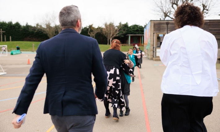 Catherine Roche and Gordon Banks are walking away from the camera towards pupils and a staff member from Churchfield Primary in their playground