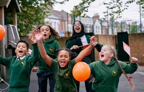 five primary school aged children outside in the playground cheering and running around with Place2Be balloons and flags