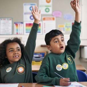 A young boy and girl with Children's Mental Health Week stickers on, sitting down in a classroom with their hands up ready to answer a question