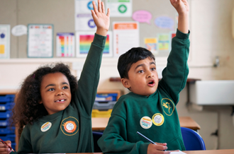Two primary age children wearing school uniform, sat in a classroom with their hands raised. Both are wearing stickers with the Children's Mental Health Week logo on.
